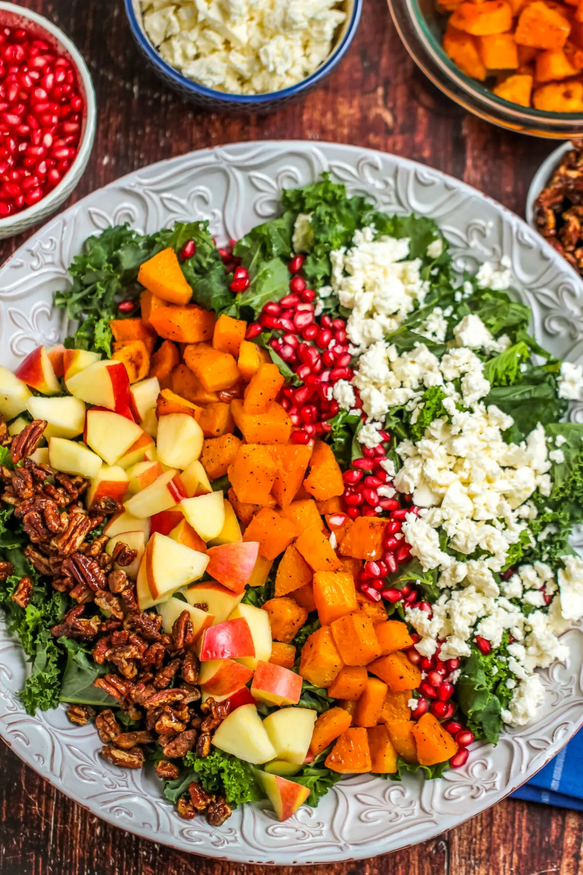 candy pecans, diced apples, roasted squash, pomegranate arils, and feta cheese on a bed of kale on a platter.